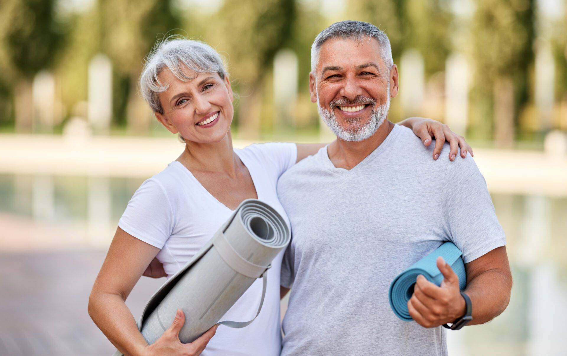 Smiling senior couple husband and wife embracing while standing at park with exercise mats